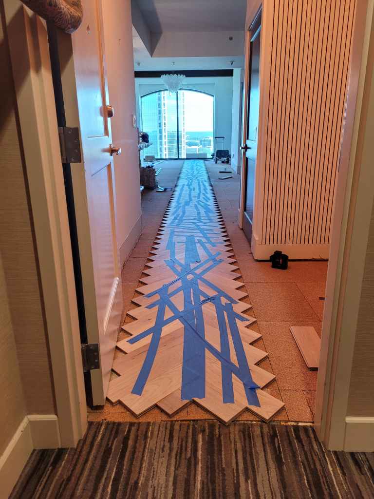 Dry-lay center line for the white oak herringbone marked with blue tape down the Buckhead high-rise hallway.