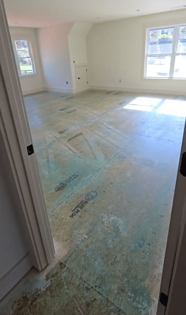Upper-level bedroom plywood subfloor in Canton, GA new-construction home after Wakol PU 280 primer application, viewed from doorway with two windows on the far wall