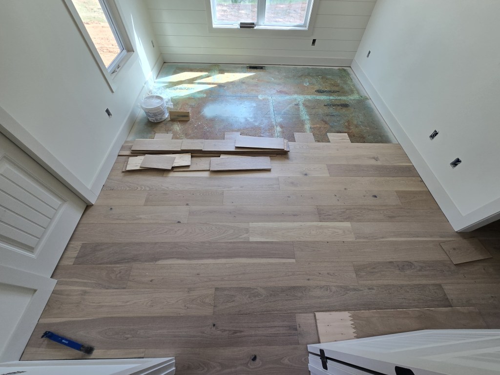 Upper-level bedroom in Canton, GA during RW Floors Kells European white oak installation, with installed planks in the foreground, loose boards staged for blending mid-room, and primed plywood subfloor at the window wall