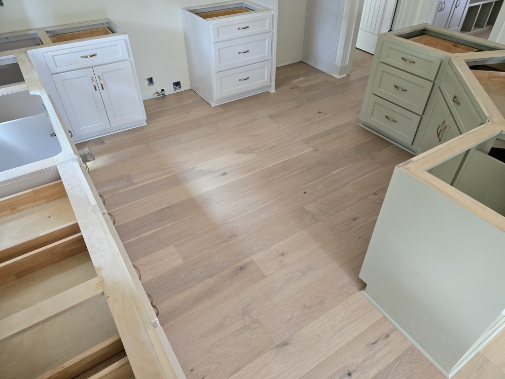 Finished RW Floors Kells European white oak flooring running through a kitchen in Canton, GA, with white perimeter cabinetry on the left, a sage green island base on the right, and cabinets still in mid-install