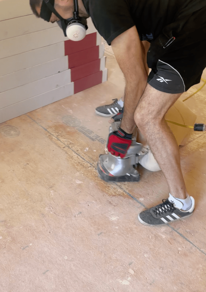 Classy Flooring ATL installer edge-sanding a plywood sheet seam during subfloor preparation in a Canton, GA home, with RW Floors Kells flooring cartons staged against the wall