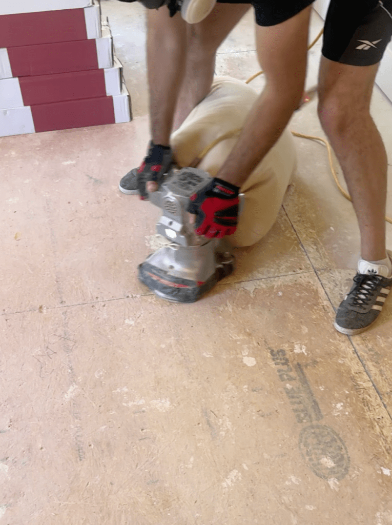 Classy Flooring ATL installer edge-sanding a plywood sheet seam during subfloor preparation in a Canton, GA home, with RW Floors Kells flooring cartons staged against the wall