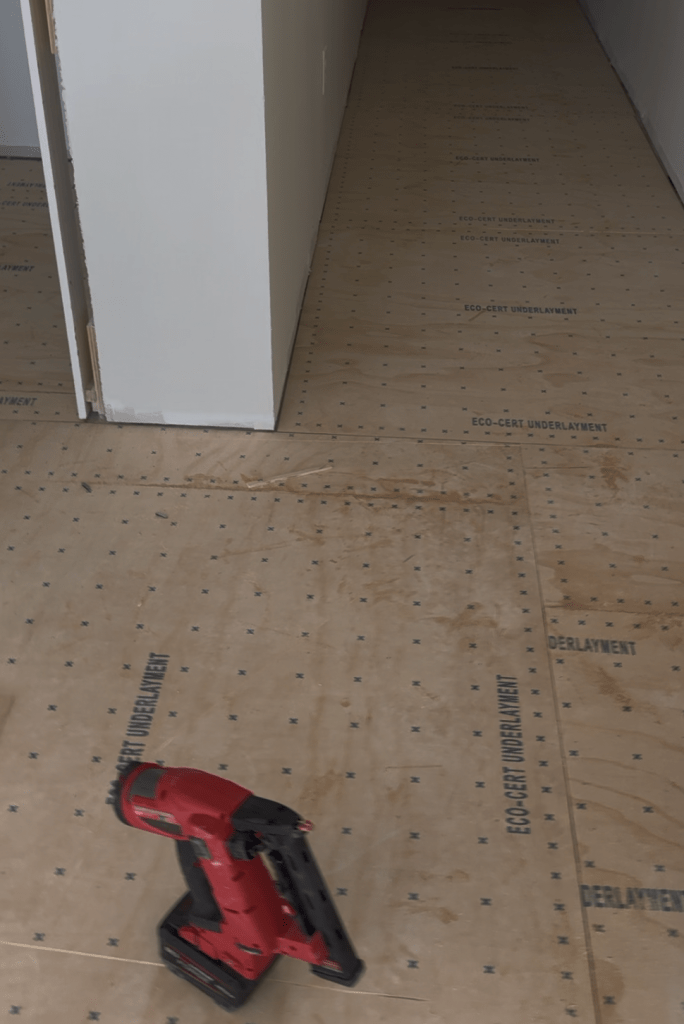 New plywood overlay fastened down a hallway with a cordless nailer resting on the panels