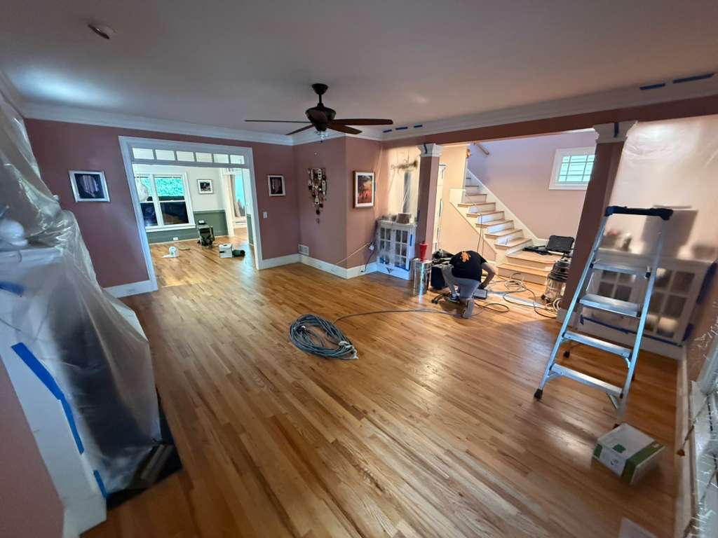 Open-plan living area with worn amber finish on red oak floors at the start of a refinishing project in Atlanta, crew preparing equipment and dust containment