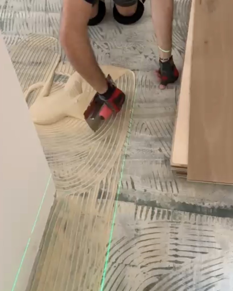 Classy Flooring ATL installer troweling urethane adhesive across the substrate along a green laser layout line, with European white oak plank staged for installation