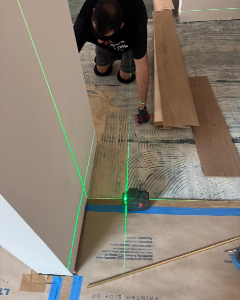 Classy Flooring ATL installer working to a green cross-line laser that projects perpendicular layout lines across the substrate, setting European white oak plank into troweled adhesive