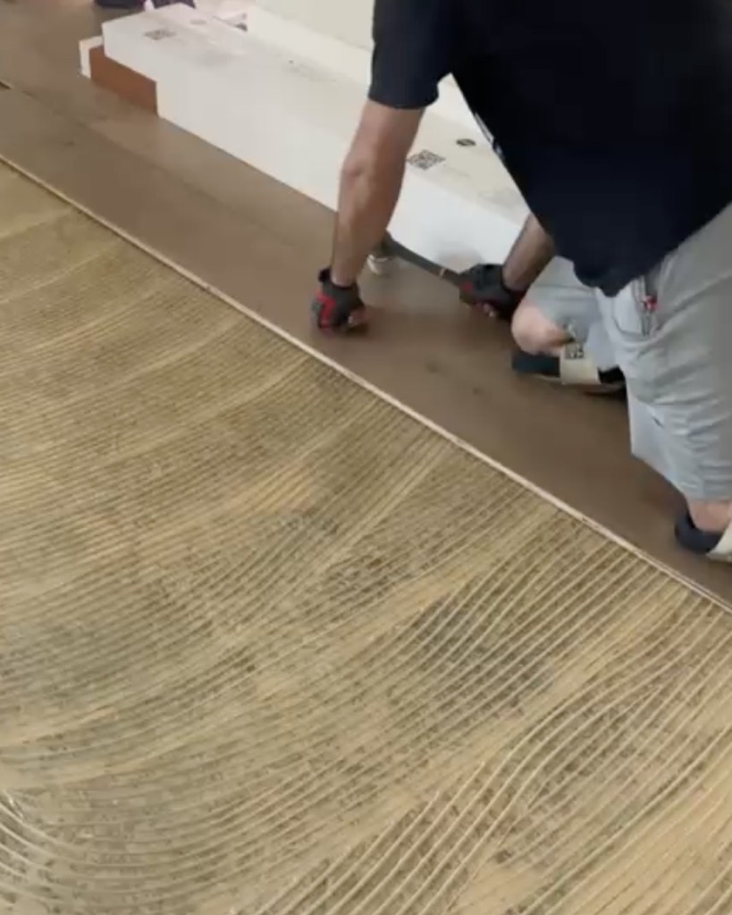 Classy Flooring ATL installer crouched setting a European white oak board into the field, with troweled adhesive ridges across the floor in front