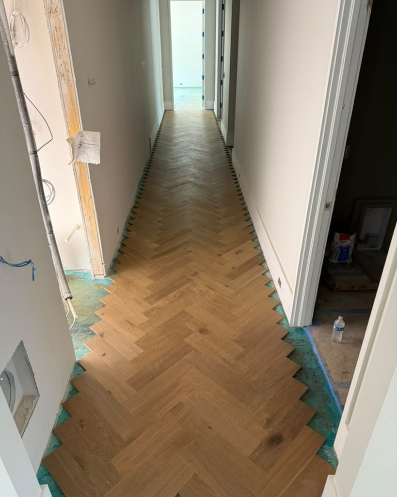 European white oak herringbone field laid down a hallway during installation, the perimeter still open over the moisture-barrier subfloor before the border and brass were set
