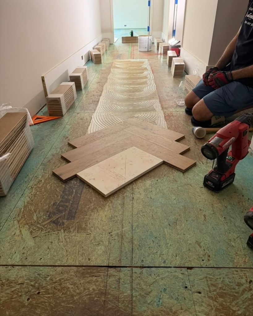 Classy Flooring ATL installer kneeling at the leading edge setting European white oak herringbone pieces into troweled adhesive, with shop-milled pieces stacked along the walls