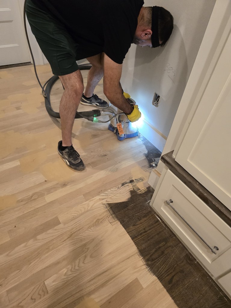 Classy Flooring ATL installer edge-sanding the perimeter of a white oak floor at a cabinet base with a handheld edger during a refinishing project in a Buckhead, Atlanta residence.