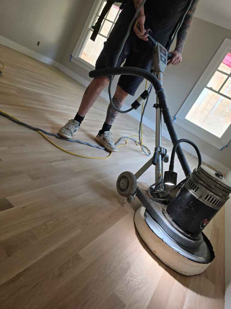 Classy Flooring ATL installer refining a bare hardwood floor with an FG Discus planetary sander in a window-lined room during a refinishing project in a Buckhead, Atlanta residence.
