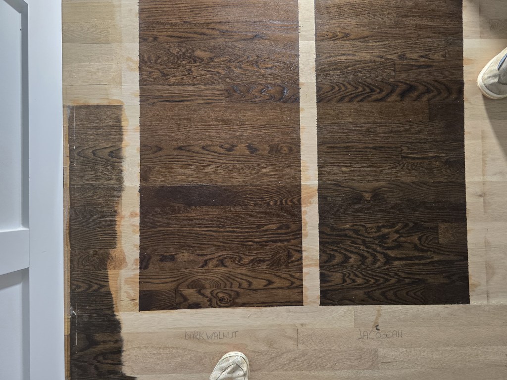 Two DuraSeal stain samples, Dark Walnut and Jacobean, brushed side by side on a bare white oak floor and hand-labeled for client comparison during a refinishing project in a Buckhead, Atlanta residence by Classy Flooring ATL.