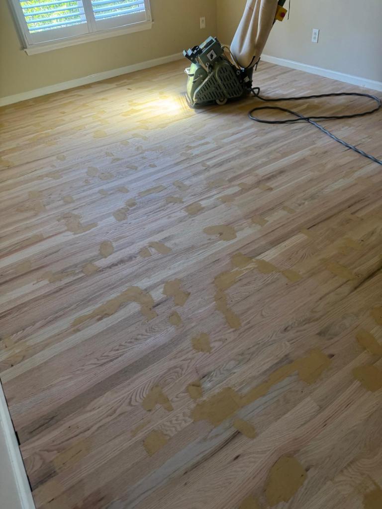 Belt sander cutting a newly installed 2¼-inch red oak strip floor during sanding, wood filler visible across the boards before final grit, Alpharetta bedroom