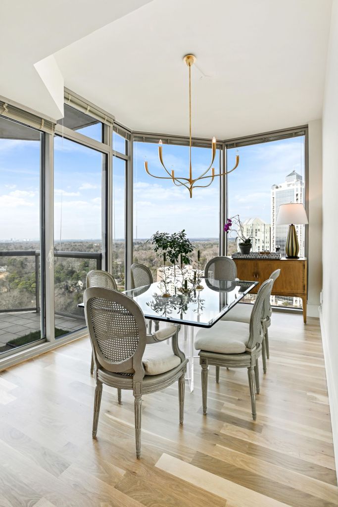 Corner dining area of a Buckhead high-rise condominium in Atlanta, with a glass table, four gray cane-back chairs and a gold branch chandelier in front of floor-to-ceiling windows on two sides overlooking the city skyline, on a 4-inch solid white oak floor installed and site-finished in LOBA 2K Duo satin by Classy Flooring ATL.