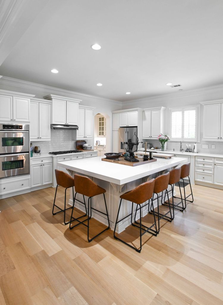 Vertical view of the kitchen in a Chateau Elan residence in Braselton, GA, with the white island and cognac leather counter stools in the center, white cabinets, a double wall oven and a window behind, on 7-inch rift and quartersawn engineered white oak installed and site-finished with Bona NordicSeal and Bona Traffic HD by Classy Flooring ATL.