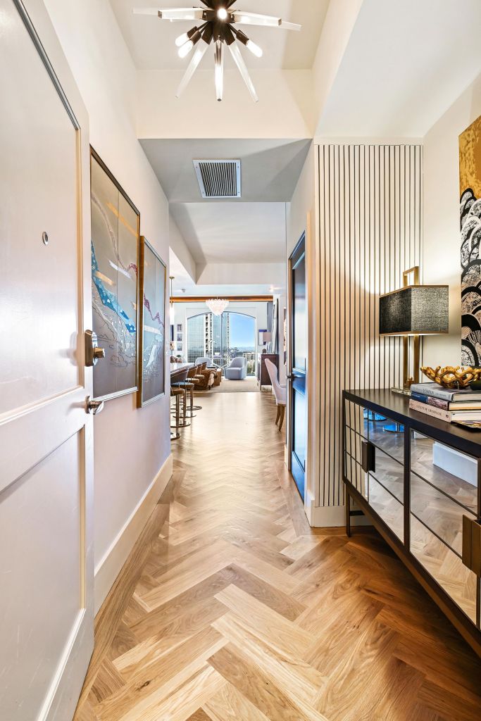 Hallway of a Buckhead high-rise condominium in Atlanta seen from the entry, with a dark door on the left, a mirrored console with a black lamp on the right, a fluted wall panel, a sputnik light and a view through to the living room windows, on a solid white oak herringbone floor running down the hallway, installed and site-finished in LOBA 2K Duo satin by Classy Flooring ATL.